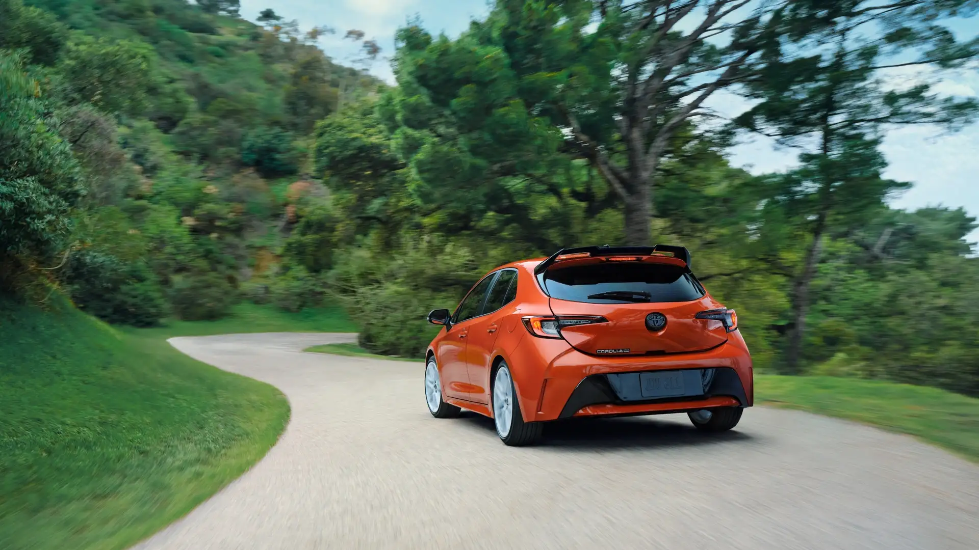 2026 Toyota Corolla Hatchback driving on a winding road, showcasing sporty exterior styling, compact hatchback design, and dynamic rear profile.