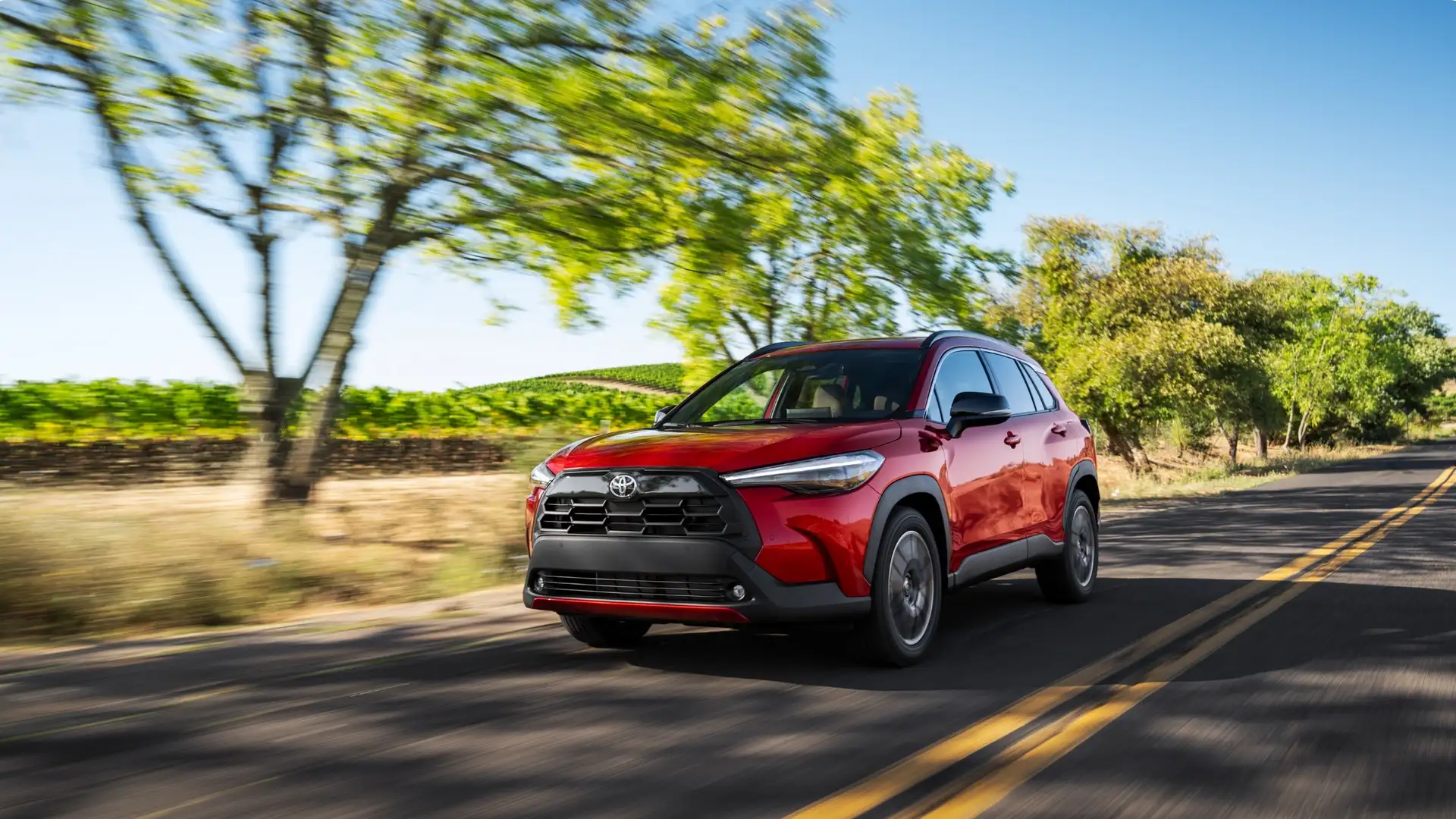 2025 Toyota Corolla Cross driving on a tree-lined road, showcasing family-friendly space and versatility for drivers in Greencastle PA.