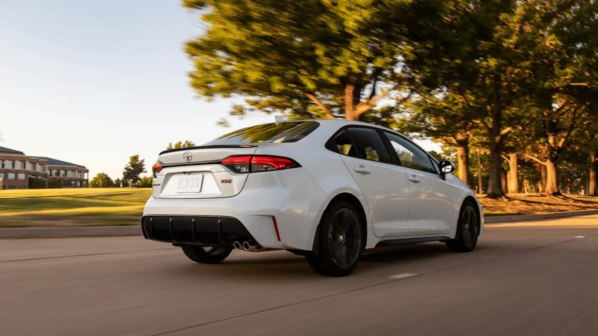 Rear three-quarter view of a white 2026 Toyota Corolla sedan driving on a city street, highlighting sleek exterior styling and LED lighting.