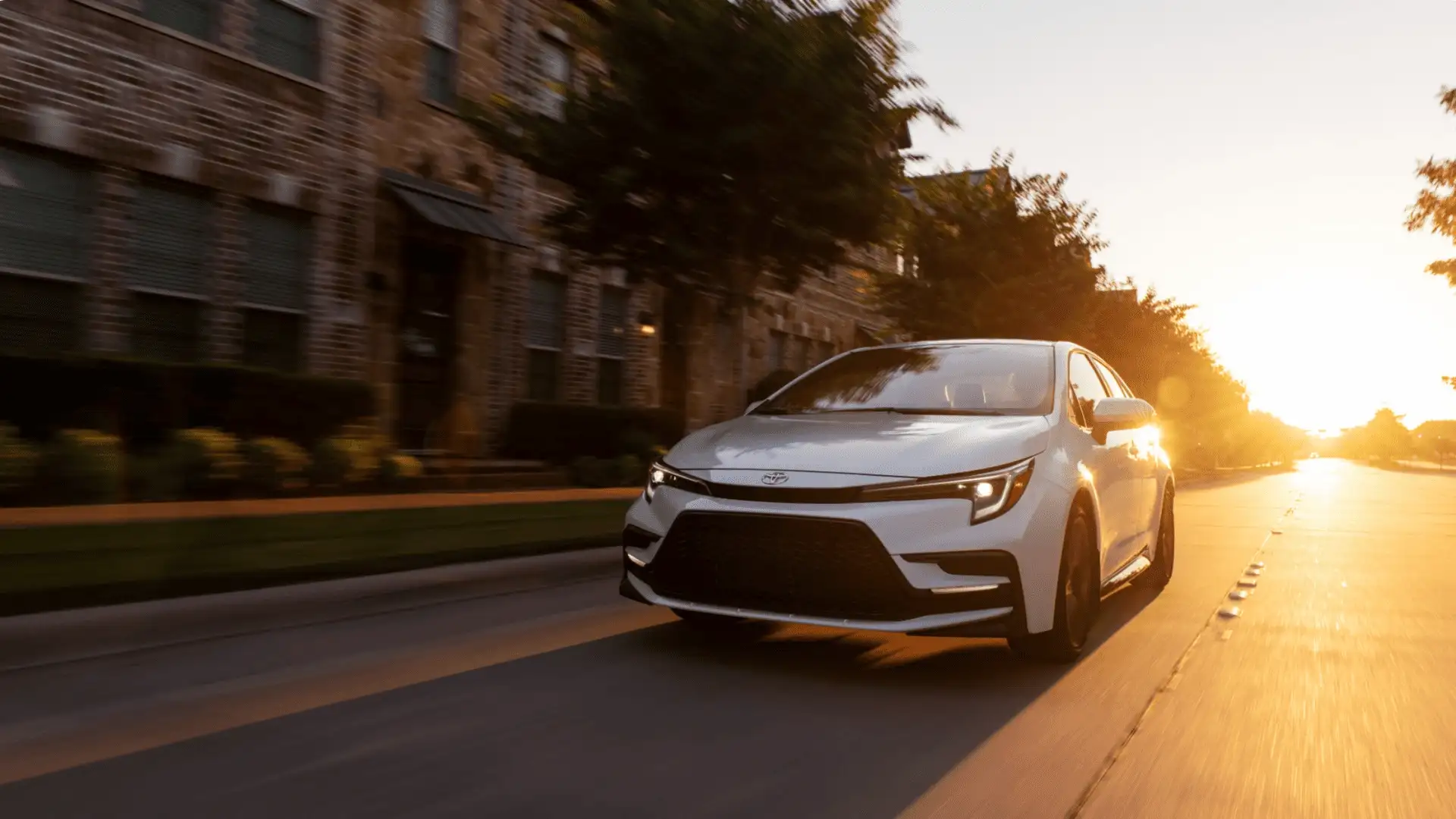 White 2026 Toyota Corolla sedan driving through a tree-lined road near Hagerstown, MD, showcasing its modern compact car design.