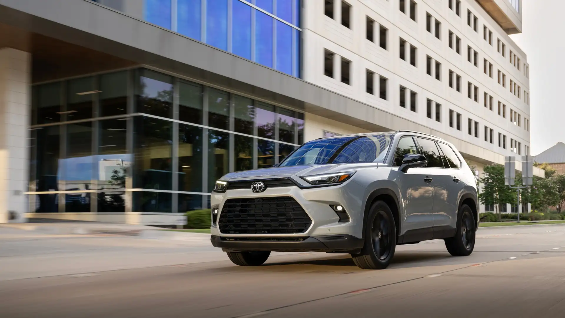 2026 Toyota Grand Highlander in silver with black wheels driving past a modern glass office building, comparing fuel efficiency near Hagerstown MD