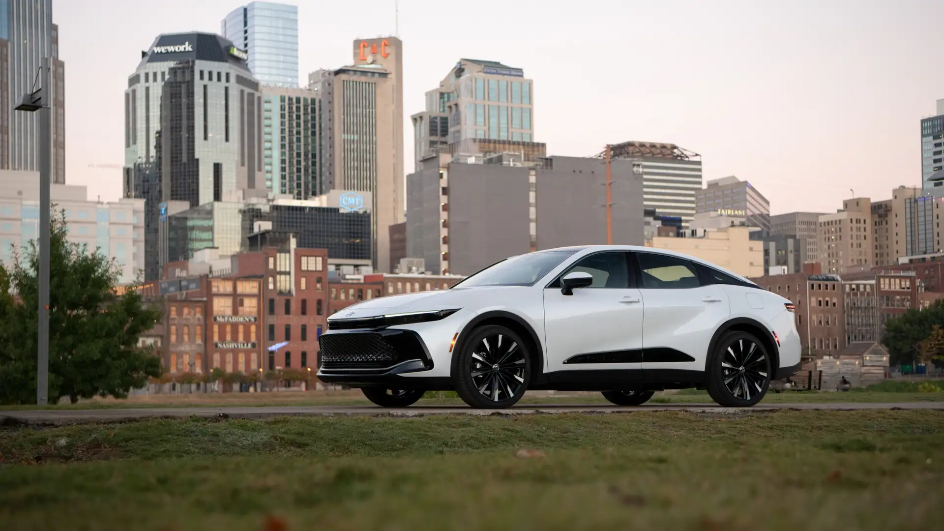 2026 Toyota Crown in white with black alloy wheels parked on a city park lawn with a downtown Nashville skyline in the background, ideal for Hagerstown MD commuters