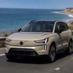 2025 Volvo EX90® electric SUV front three-quarter driving view in beige on coastal highway