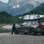 2025 Volvo XC60 Plug-In Hybrid SUV with roof-mounted kayak parked by alpine lake, rear three-quarter view in gray