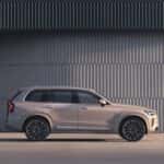 2025 Volvo XC90 luxury SUV shown in full side profile against modern architectural backdrop, metallic beige finish
