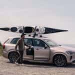 2025 Volvo XC90 Plug-In Hybrid SUV with kayak on roof at coastal overlook, driver loading gear, side view in silver